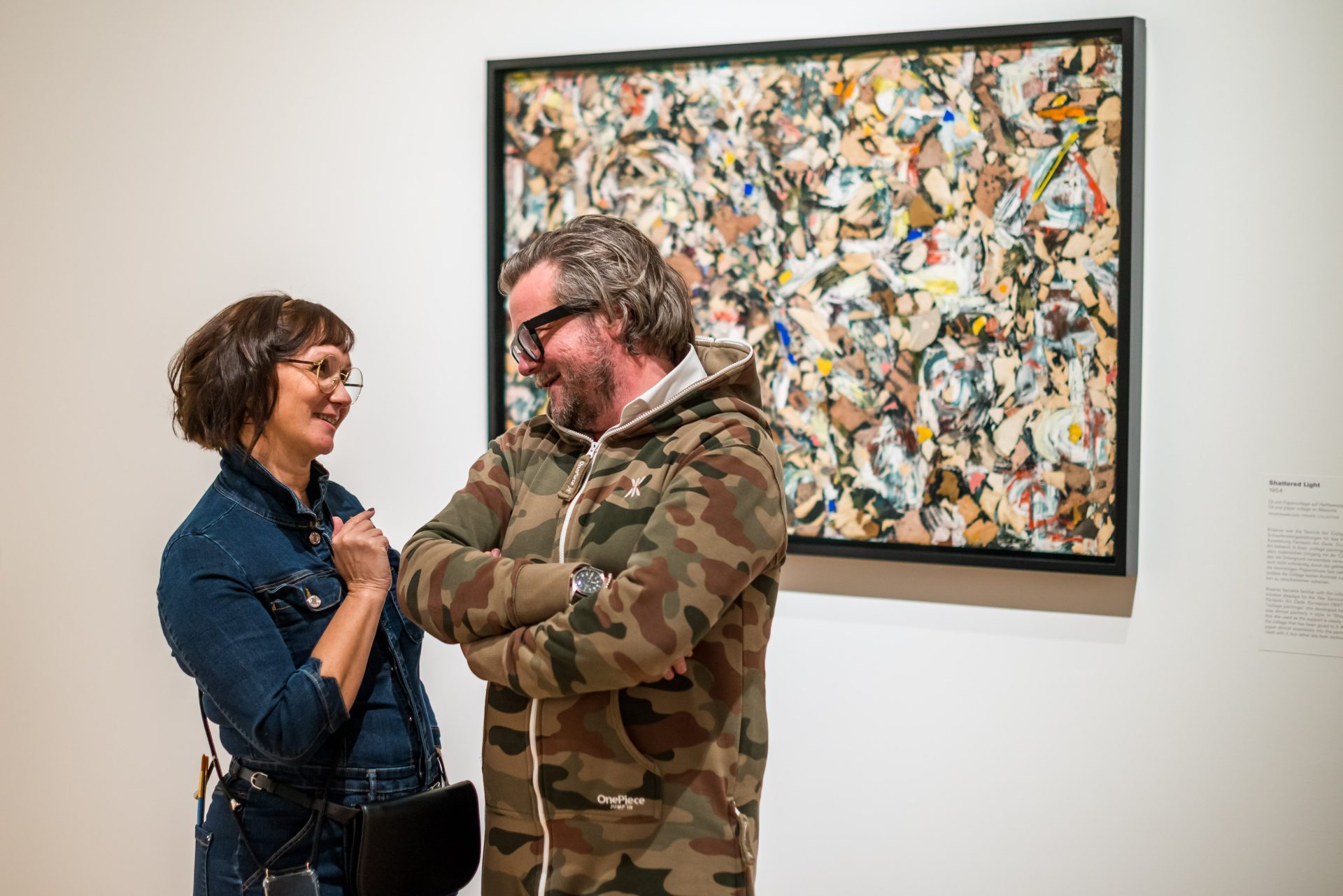 Zwei Personen lächeln vor einem bunten Kunstwerk in einer Galerie.