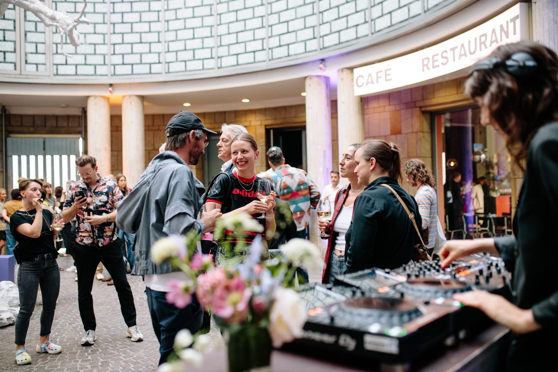 Menschen feiern in einem Café, Musik erklingt, und Blumenarrangements sowie eine DJ-Konsole sind zu sehen.