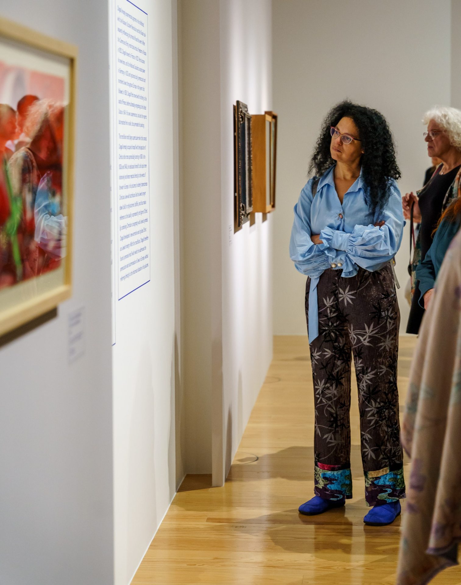 Zwei Frauen im Museum betrachten Wandkunst. Eine trägt eine blaue Bluse und bunte Hosen.