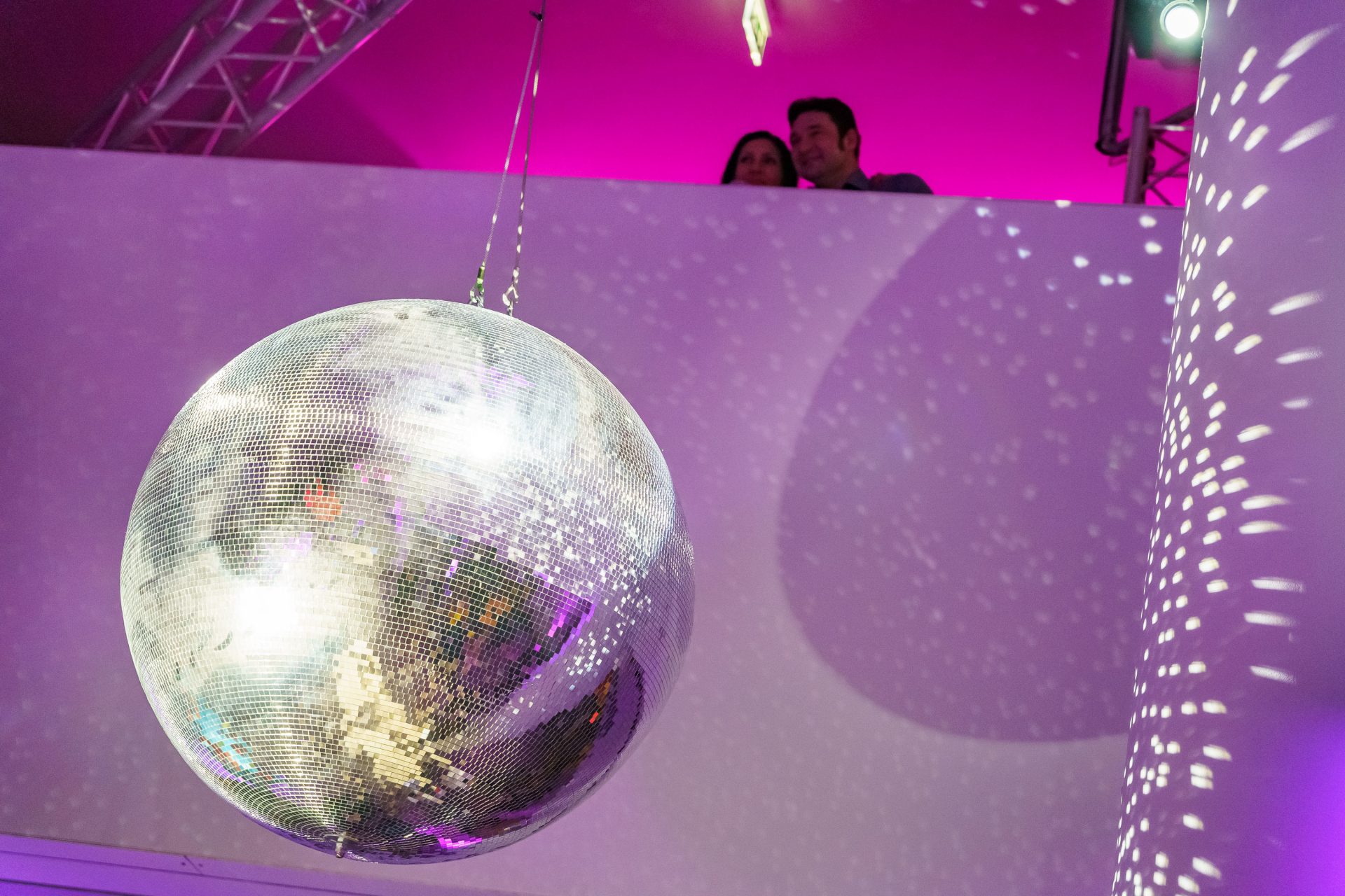 Diskokugel hängt von der Decke, farbige Lichter spiegeln sich an den Wänden. Ein Paar sieht von oben zu.
