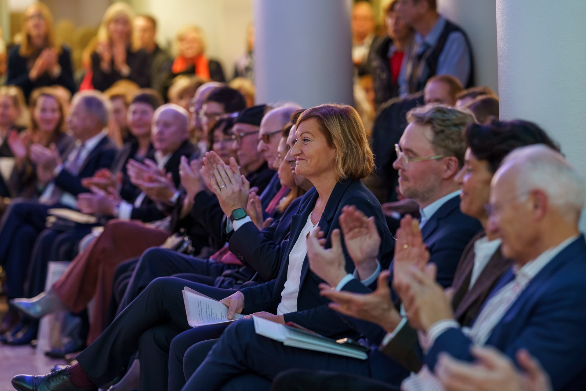 Menschen applaudieren im Publikum, einige lächeln, während sie an einer Veranstaltung teilnehmen.