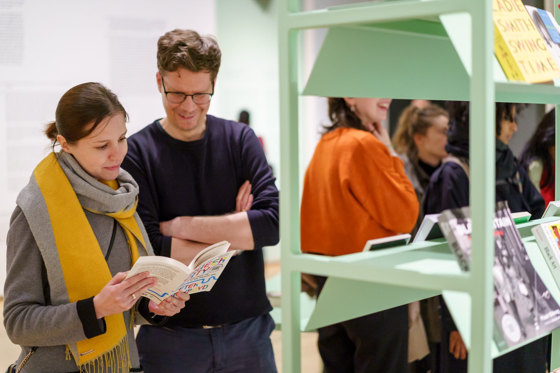 Zwei Personen in einer Ausstellung betrachten ein Buch, während im Hintergrund weitere Besucher zu sehen sind.