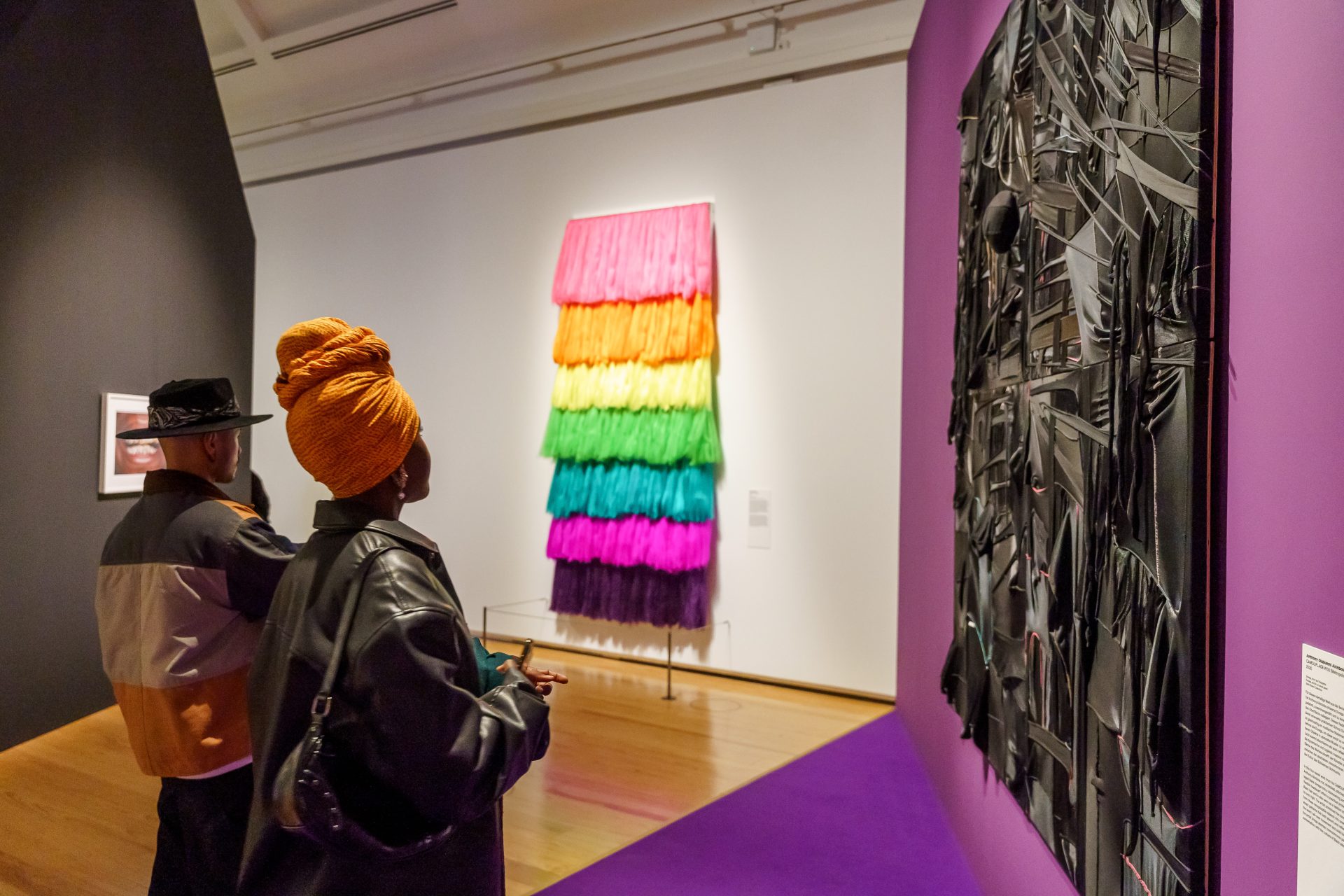 Zwei Personen betrachten in einer Galerie bunte Kunstwerke, darunter ein gestreiftes Werk und eine dunkle, strukturierte Wand.