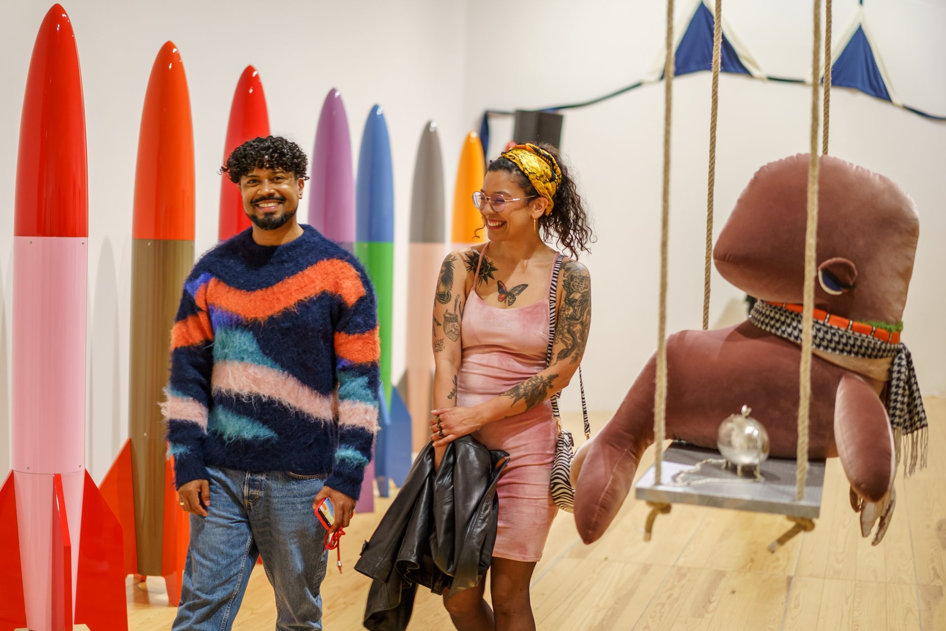 Zwei Personen stehen in einer Galerie mit bunten Skulpturen und einem großen, schwingenden, weichen Objekt.