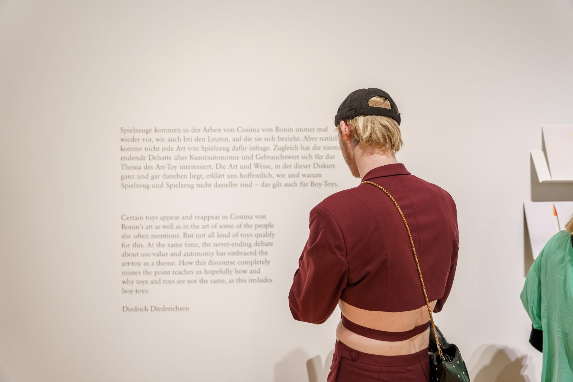 Person in rotem Anzug und Kappe steht vor einer Wand mit Text und betrachtet eine Ausstellung.