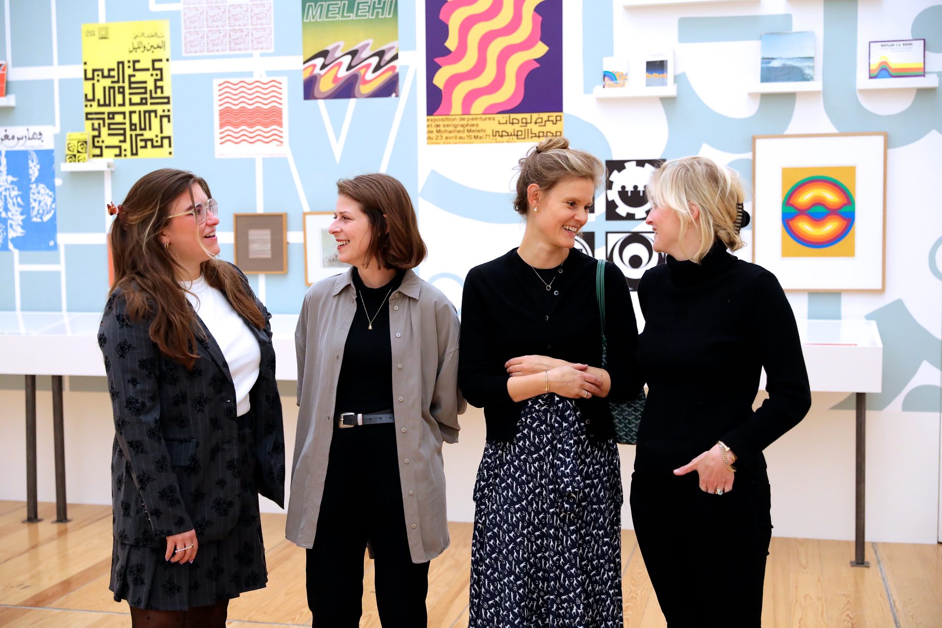 Vier Frauen stehen in einem Museum, umgeben von bunten Kunstwerken. Sie lächeln und führen ein Gespräch.
