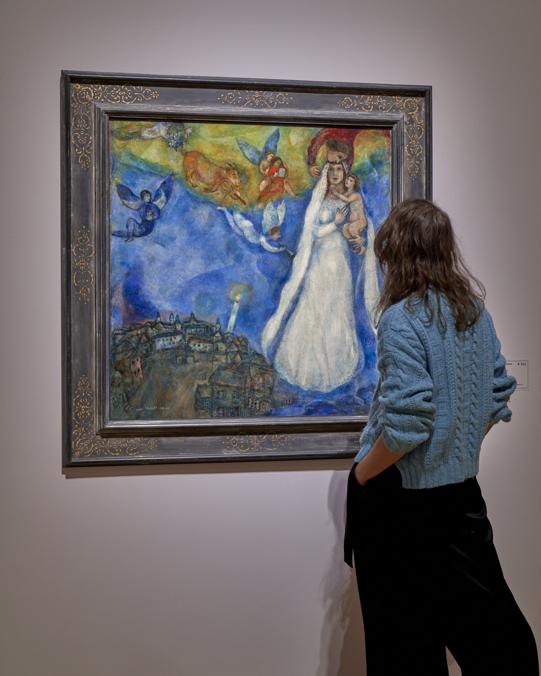 Eine in Weiß gekleidete Frau betrachtet ein farbenfrohes Gemälde von Engeln und einer Stadt im Hintergrund.