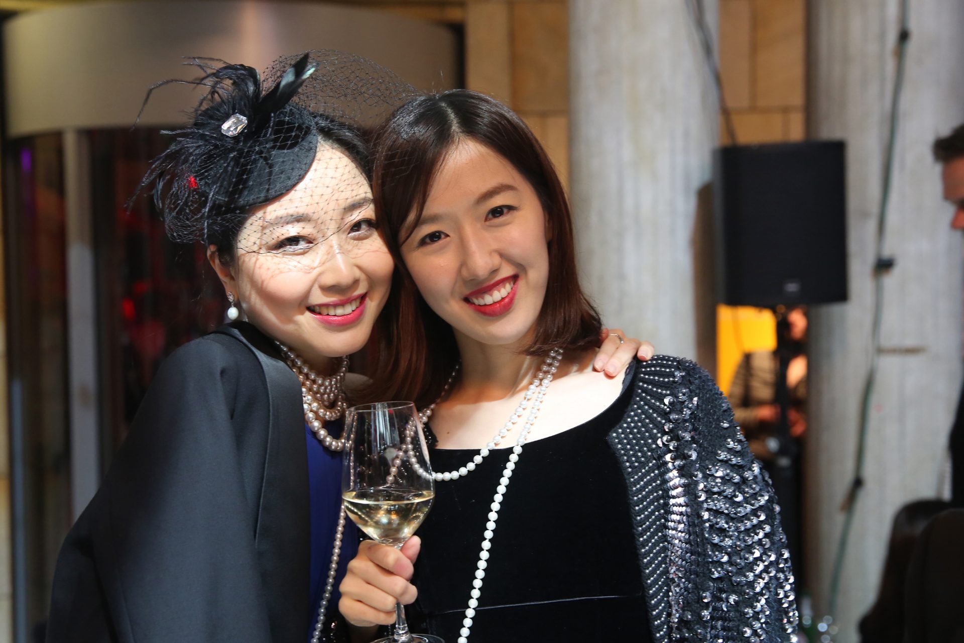 Zwei Damen in eleganter Abendmode lächeln, eine hält ein Glas Sekt, im Hintergrund herrscht festliche Stimmung.