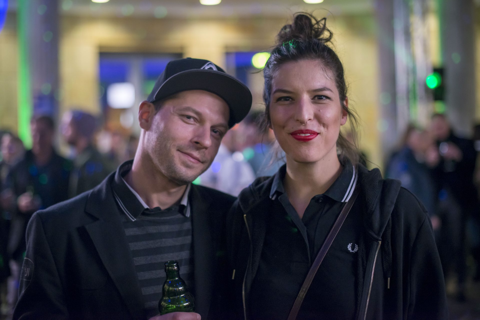 Ein Paar lächelt auf einer lebhaften Veranstaltung, im Hintergrund unscharfe Personen und bunte Lichter.