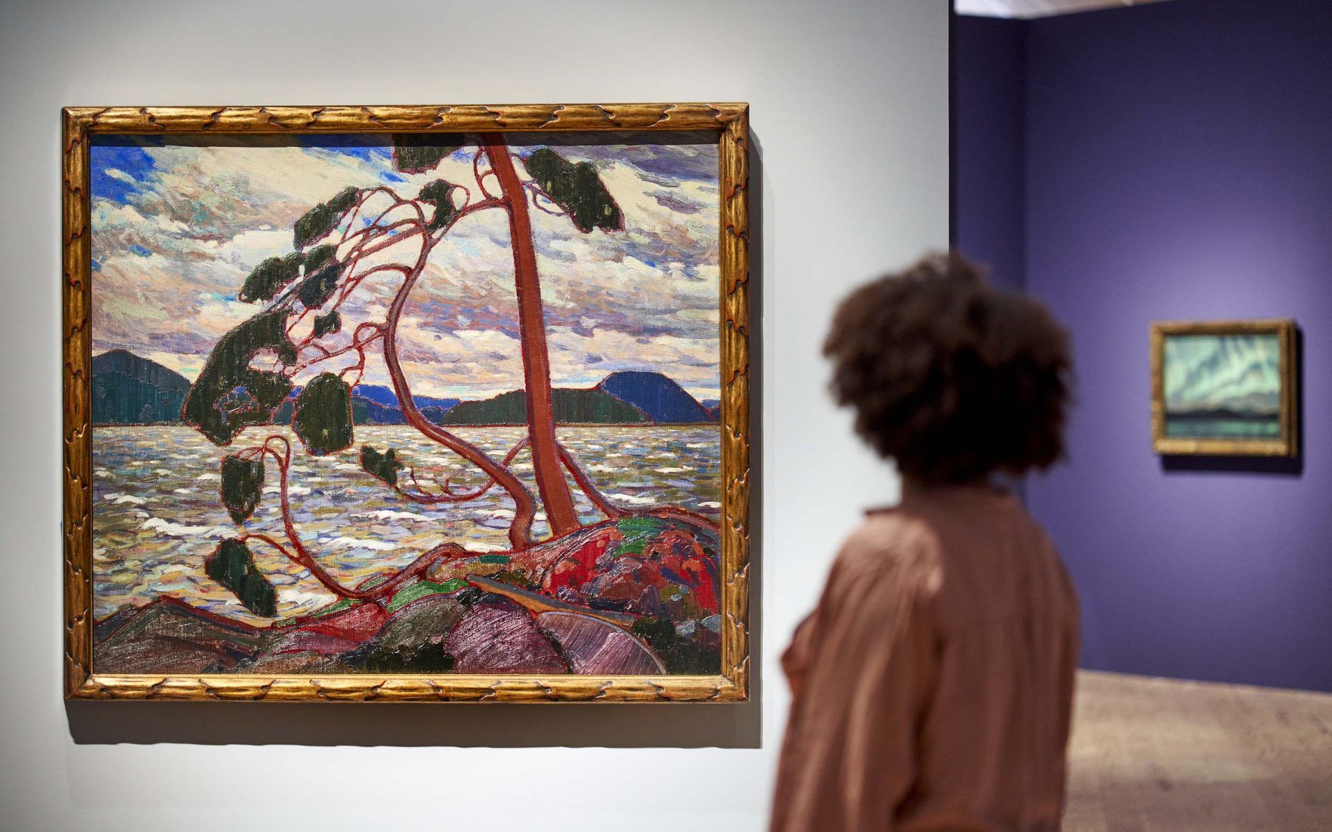 Person betrachtet ein lebhaftes Landschaftsgemälde mit Bäumen und Wasser in einer Galerie.