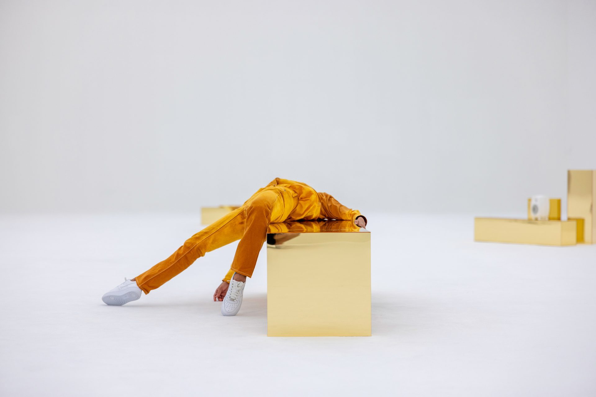 Person in gelbem Outfit lehnt entspannt auf einem goldenen Block in einer minimalistischen, hellen Umgebung.