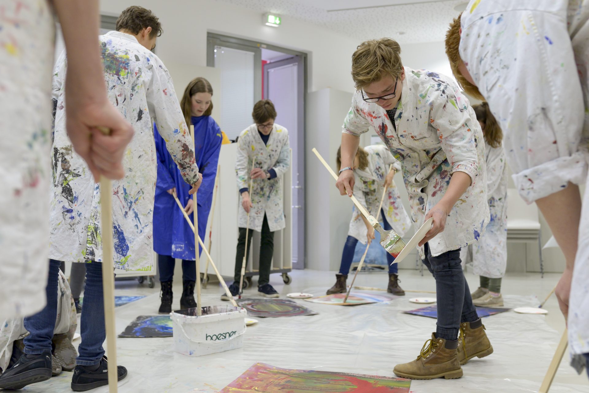 Gruppe von Menschen in weißen Maleranzügen, die auf dem Boden mit langen Pinseln kreativ arbeiten.