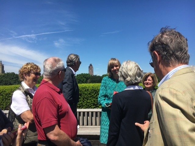 Gruppe von Menschen bei sonnigem Wetter im Freien, im Gespräch vor einer grünen Hecke und Gebäuden im Hintergrund.