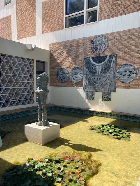 Skulptur in einem Teich mit Wasserlilien, neben einer Wand mit Reliefs und Mosaiken. Architektur im Hintergrund.