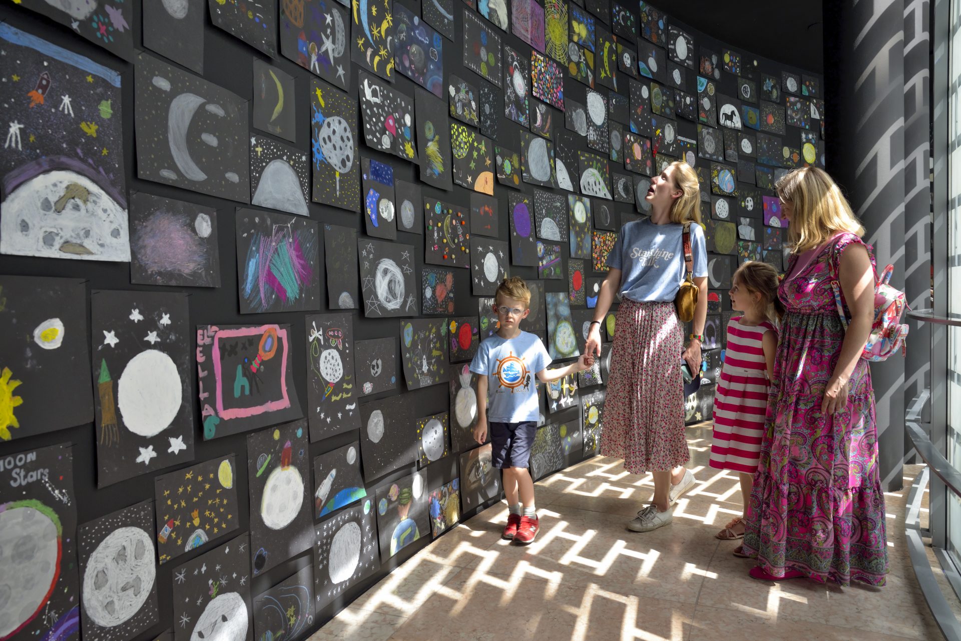 Familie betrachtet im Ausstellungsraum eine Wand mit bunten Kinderzeichnungen zu astronomischen Themen.