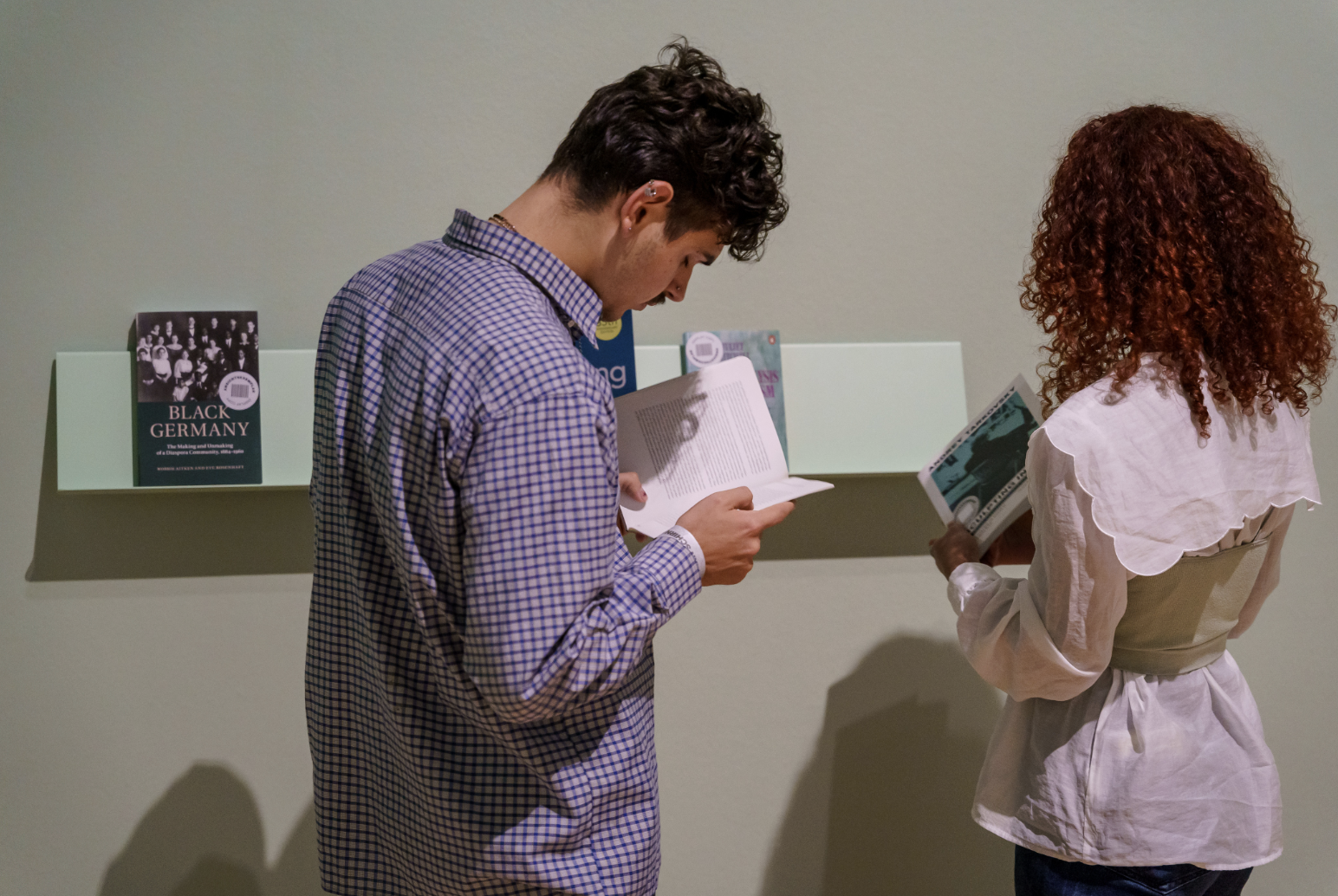 Zwei Personen lesen Bücher in einer Galerie mit Regalen im Hintergrund.