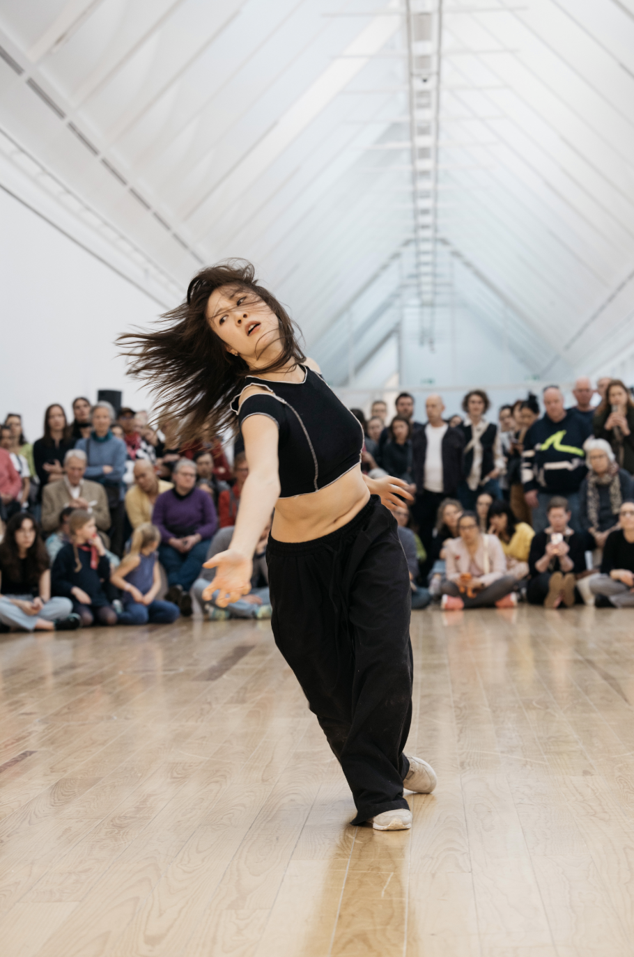 Tänzerin in lässiger Kleidung, die im Zentrum einer Zuschauermenge in einem hellen, modernen Raum tanzt.