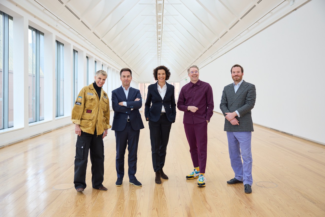 Fünf Personen in eleganter Kleidung stehen in einem modernen, lichtdurchfluteten Raum mit Holzfußboden.