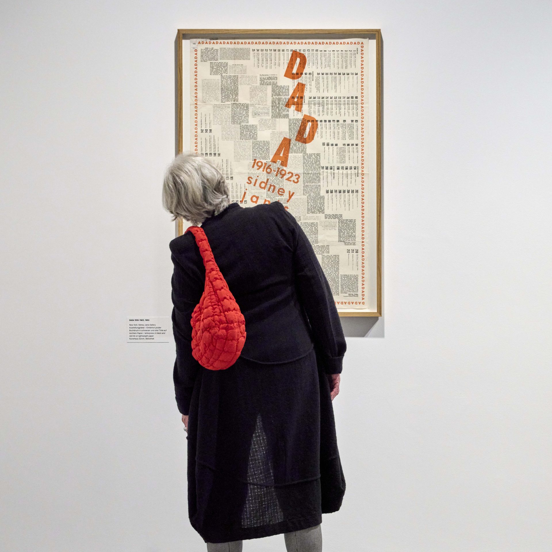 Eine Person mit roter Tasche und schwarzem Mantel betrachtet ein Dada-Kunstwerk in einer Galerie.