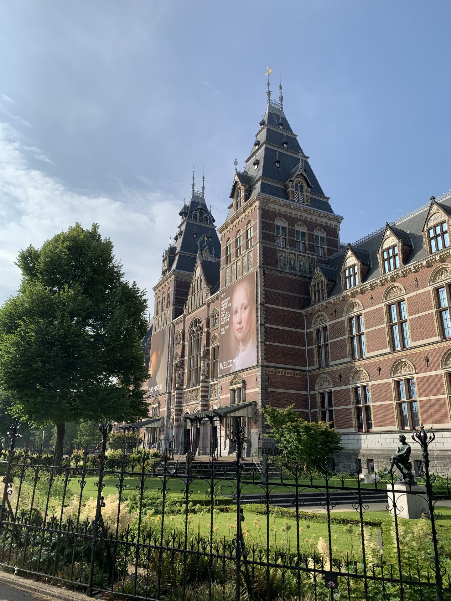 Historisches Gebäude mit Türmen, grünem Garten und Kunstplakaten bei blauem Himmel und sonniger Atmosphäre.