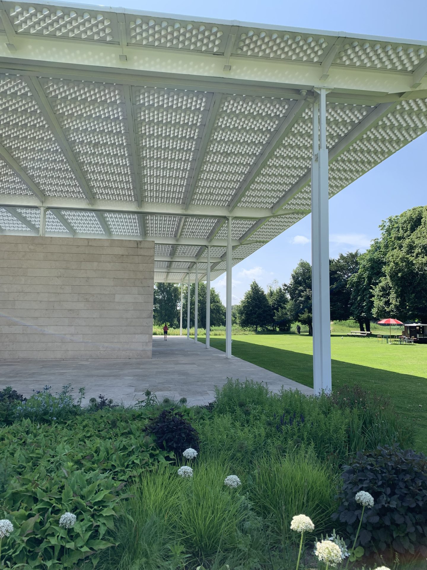 Moderne Architektur mit schattenspendendem Dach, umgeben von grünen Wiesen und blühenden Pflanzen im Freien.